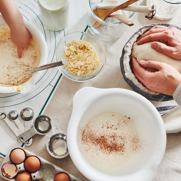 A baking table covered in bowls, ingredients and utensils with a child’s hand in one bowl and an adult’s hands in another.