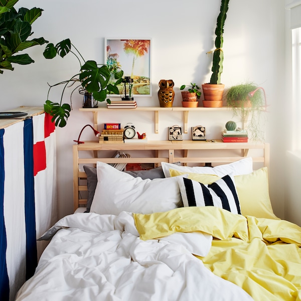 A bed in a small room with yellow and white bed linen and a pine shelving unit behind it holding plants and small objects.