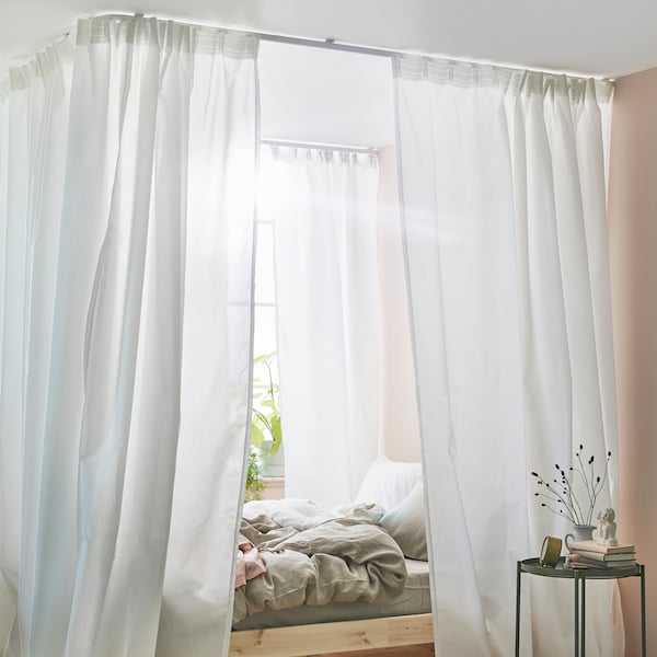 A bed stands near a window in a sunny bedroom. The bed has a canopy made using white curtains and VIDGA curtain tracks.