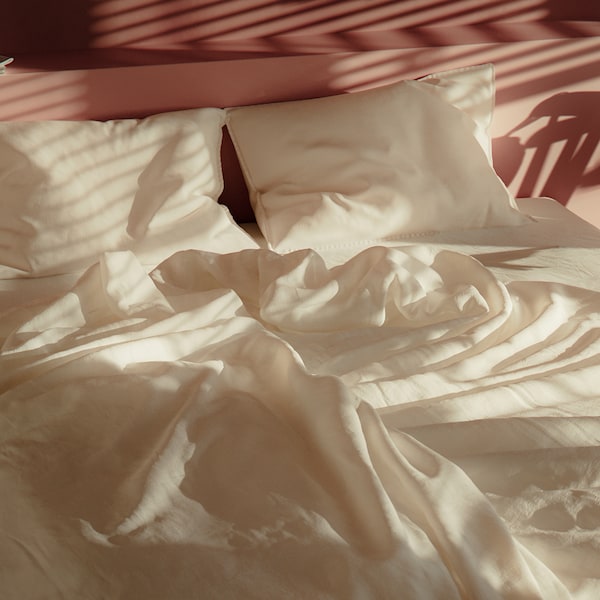 A bed with a duvet and pillows in white DYTÅG bed linen standing in a bedroom with shadows on the wall and on the bed.