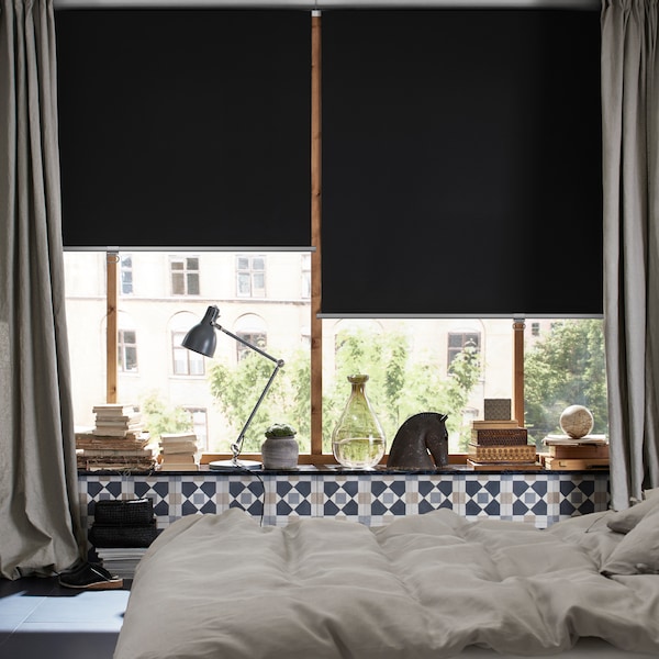 A bedroom with a bed and windows with long curtains and TRETUR block-out roller blinds pulled half-down.