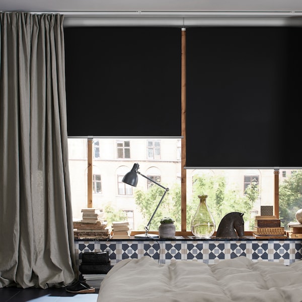 A bedroom with a bed in the middle and windows with TRETUR block-out roller blinds pulled half-down and a curtain.