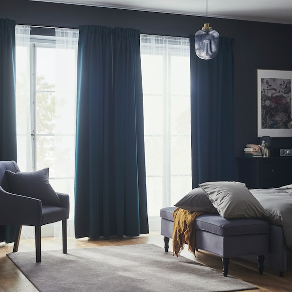 A bedroom with dark turquoise MAJGULL block-out curtains and net curtains in large windows near a bed and a bench.
