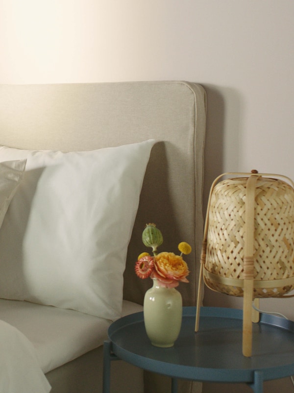 A beige upholstered bed covered in white bed linen, with a round blue table beside it that has a rattan table lamp on it.