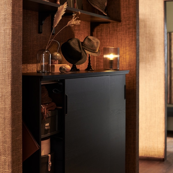 A black GALANT cabinet with one sliding door open in a cosy alcove. Hats on stands are on the top between a lamp and a vase.