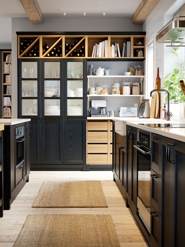 A black kitchen with a wooden floor, the edge of a black kitchen island, black cabinets and open shelves with various items.