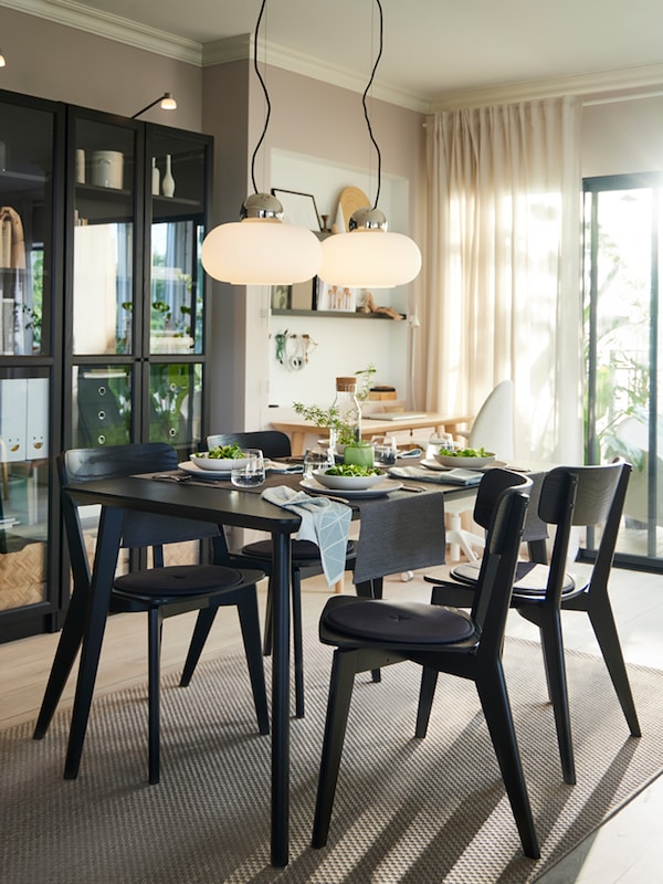A black LISABO dining table with four chairs around it laid with plates, bowls and glasses for a meal, in a bright room.