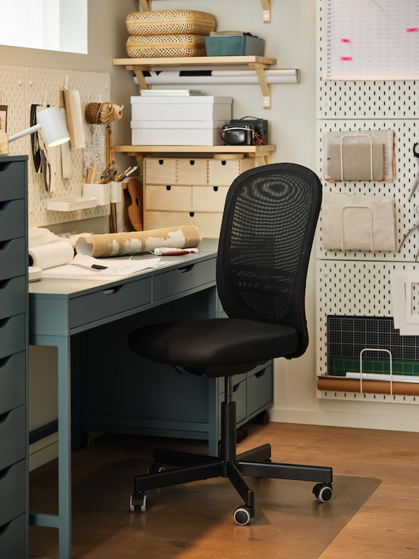 A black office chair, a floor protector, a grey-turquoise desk, white pegboards, rolls of paper.