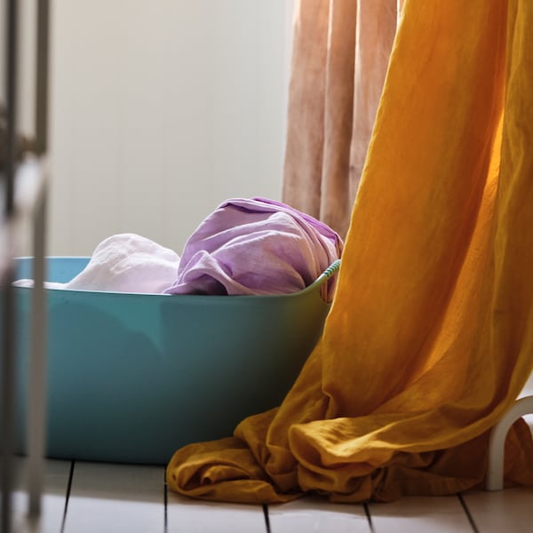 A blue TORKIS laundry basket stands on the floor next to colourful curtains.