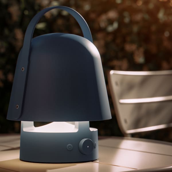 A blue VAPPEBY Bluetooth speaker lamp stands on a grey SUNDSÖ table on a terrace in the evening. The lamp is on.