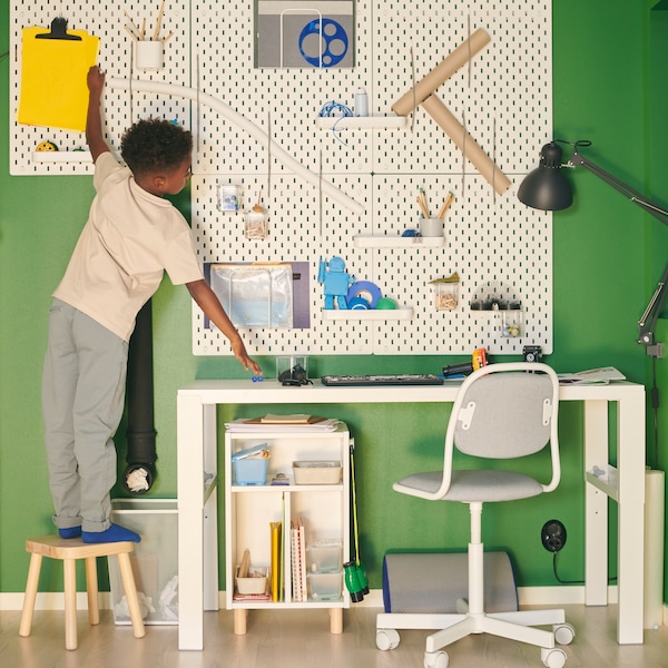 A boy standing on a stool tends to a SKÅDIS pegboard combination hung over a white PÅHL desk in a green room.