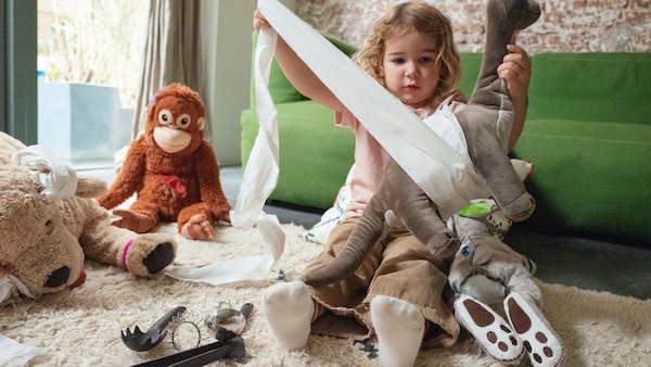 A child sits on a rug with some kitchen utensils and soft toys and wraps toilet paper around a JÄTTELIK soft toy.