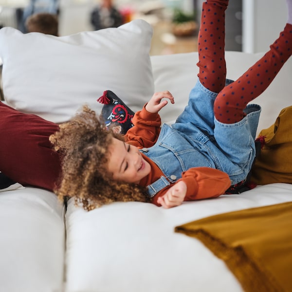 A girl happily rolls around, feet in the air, amid colorful textiles on a white GRÖNLID four-seat sofa with chaise lounge.