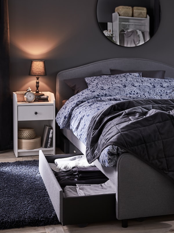 A grey upholstered bed stands in a bedroom beside a white HAUGA bedside table holding a small table lamp.