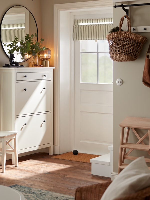 A hallway where a white HEMNES shoe cabinet stands against the wall under a black LINDBYN round mirror next to the door.