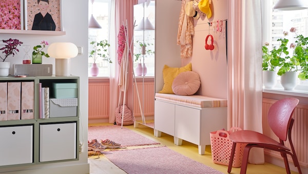 A hallway with a mirrored wardrobe, a bench with pink and yellow cushions, two rugs and hanging hats and scarves.