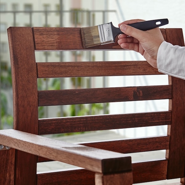 A hand holding a brush and applying VÅRDA wood stain to a wooden armchair.