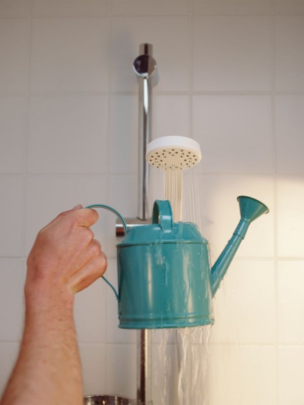 A hand holds a small blue watering can under a running shower.