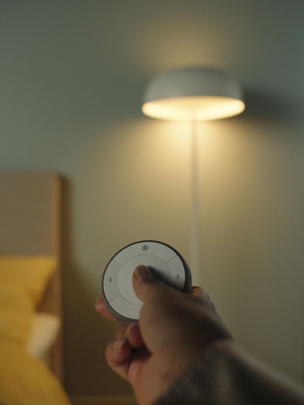 A hand on a TRÅDFRI remote control device being held out pointed towards a floor lamp standing in a room.
