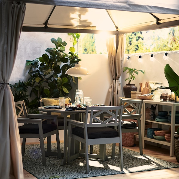 A HIMMELSÖ gazebo on a sunny and leafy garden terrace with a grey BONDHOLMEN table and chairs under it set for a cosy meal.
