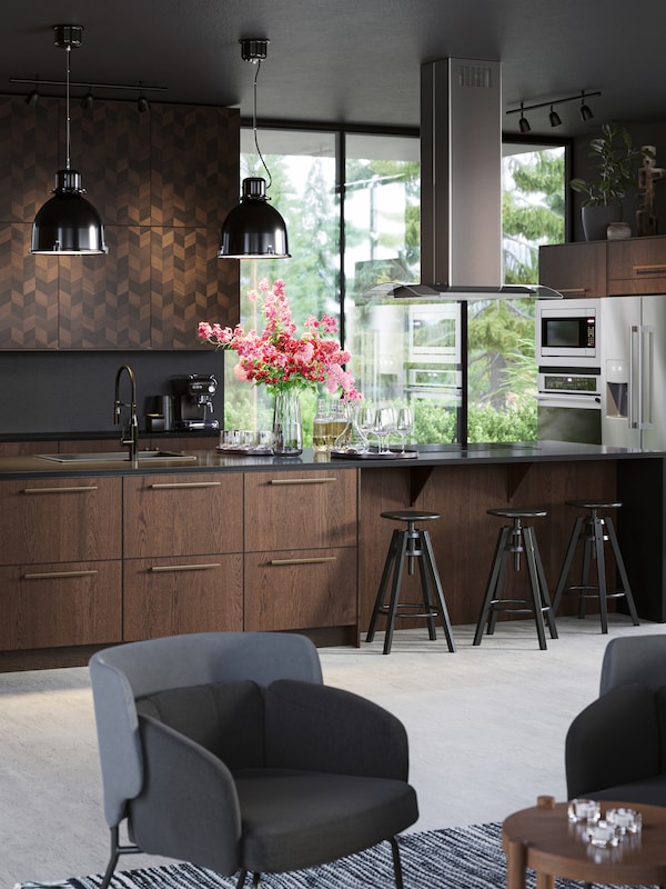 A large stylish kitchen island with wooden fronts. Black bar stools, black pendant lamps, a ceiling-mounted extractor hood.
