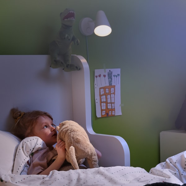 A little girl lies in bed and whispers to her favorite soft toy. A FUBBLA wall lamp attached to the wall brightens her room.