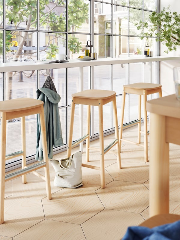A long bar with a coat on a hook underneath and birch bar stools by a floor-to-ceiling window of a restaurant.