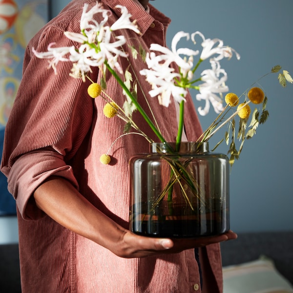 A man holding a brown KONSTFULL vase in glass with some white and yellow flowers against the backdrop of a living room.