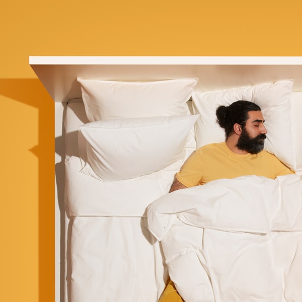 A man wearing yellow clothes sleeps in a white MALM bed with white bedding which stands on a dark yellow floor.