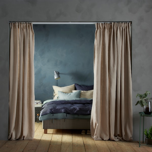 A pair of beige AINA curtains hang in a doorway leading to a bedroom with a bed with a wall lamp above it.