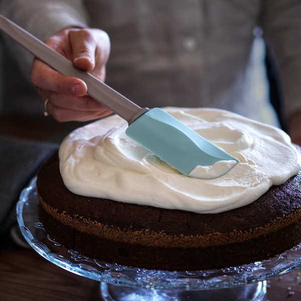 A person icing a chocolate cake on an ARV BRÖLLOP serving stand in clear glass with a BAKGLAD spatula.