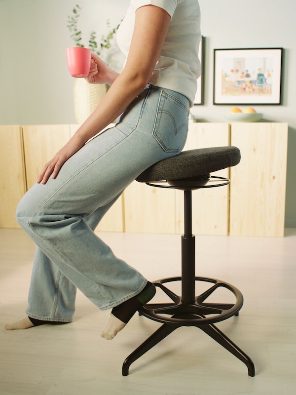 A person in blue jeans and white T-shirt resting on a LIDKULLEN active sit/stand support.