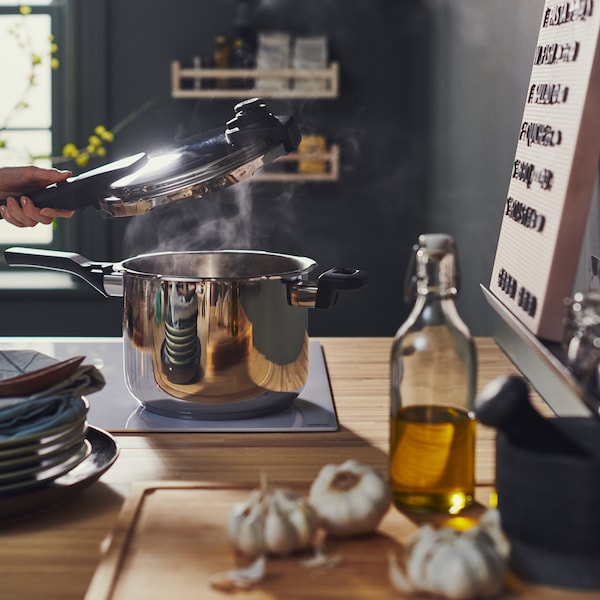 A person lifts the lid off a stainless steel IKEA 365+ pressure cooker on a hob, beside a bamboo APTITLIG butcher’s block.
