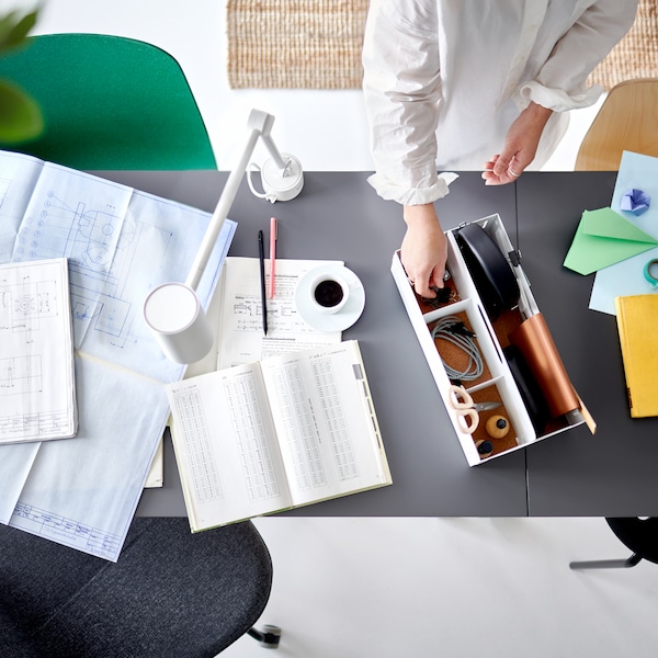 A person putting things into a white KVISSLE desk organizer that’s on top of a grey desk beside papers and a work lamp.