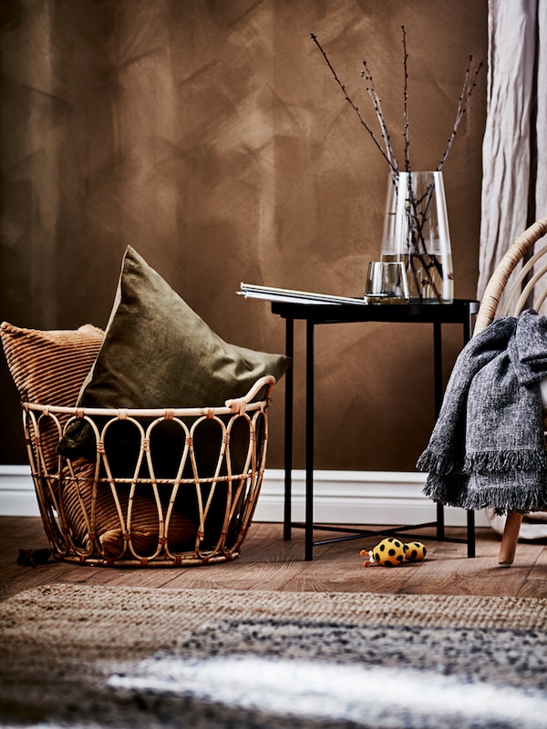 A rustic room with a SNIDAD rattan basket with warm-toned cushions in it next to a black tray table and a rattan armchair.