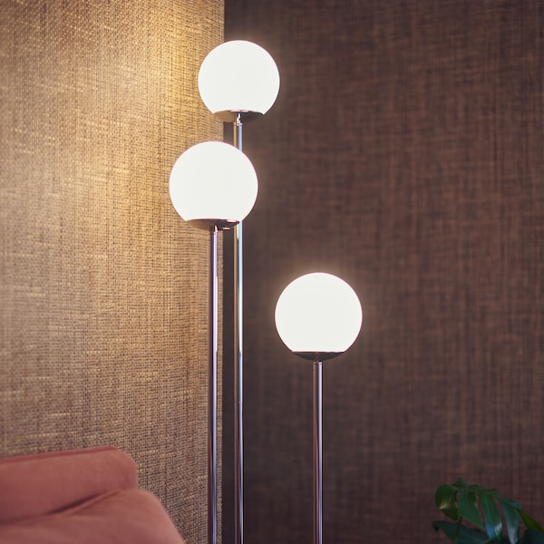 A SIMRISHAMN floor lamp with three bulbs giving off a warm glow against natural brown walls in the corner of a living room.