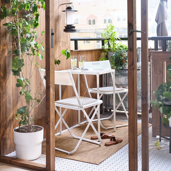 A small balcony with a white outdoor table and two white foldable chairs, a flatwoven rug and plants in plant pots.