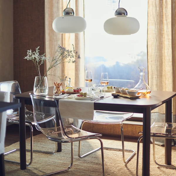 A STRANDTORP extendable table by a big, bright window set for a meal for two with food, glasses and a vase of flowers on top.