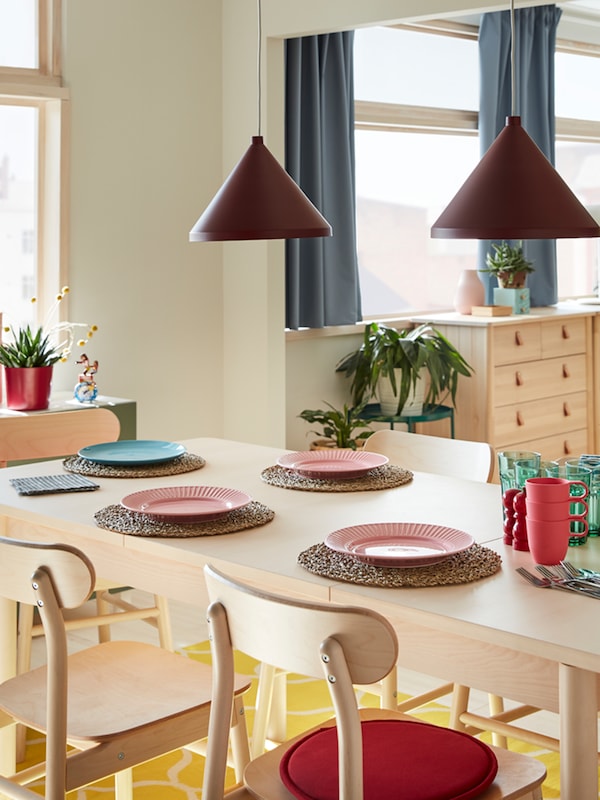 A table and chairs in birch, a yellow rug, plates in pink and blue, green glasses, cutlery and two dark red pendant lamps.