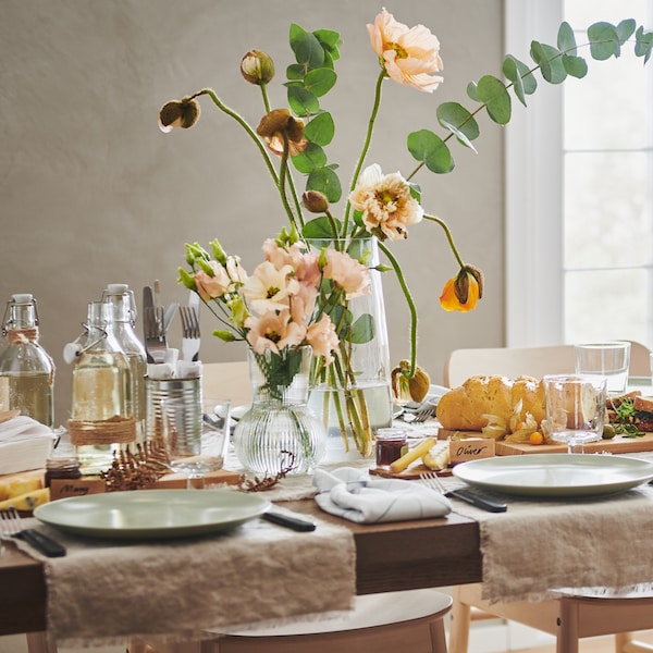 A table set for a meal with neutral tones and light green tableware. Two vases of flowers, food and bottles are on the table.