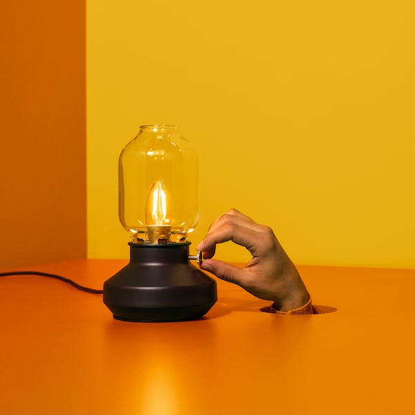 A TÄRNABY table lamp sits in a room with an orange floor. A hand pokes out through a hole in the floor and adjusts the lamp.