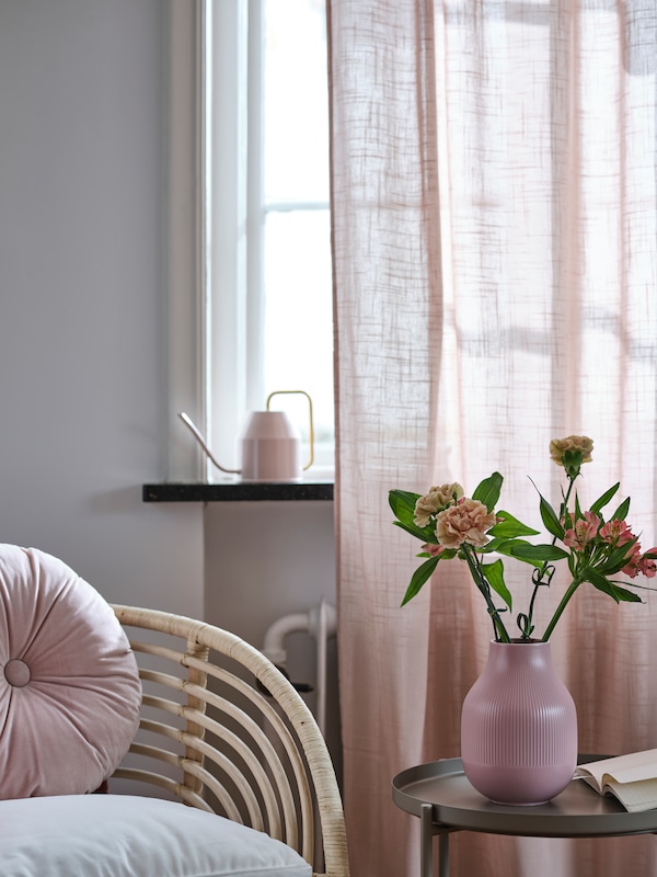 A VATTENKRASSE watering can sits in a window with SILVERLÖNN sheer curtains near a chair and a table with flowers on it.