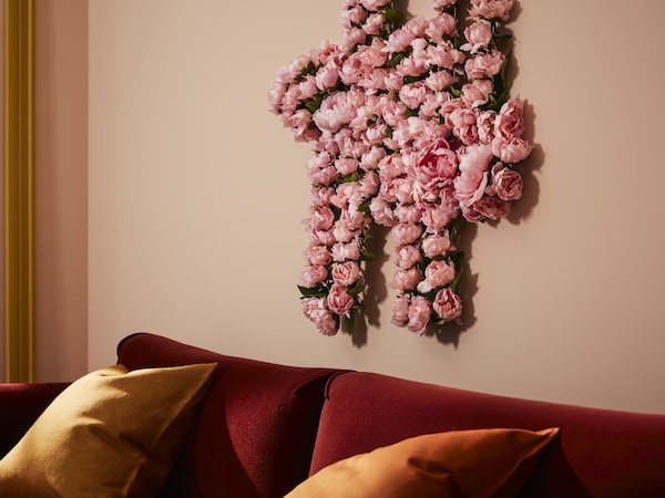 A wall decoration created from pink artificial flowers SMYCKA above the red velver sofa with yellow cushions.