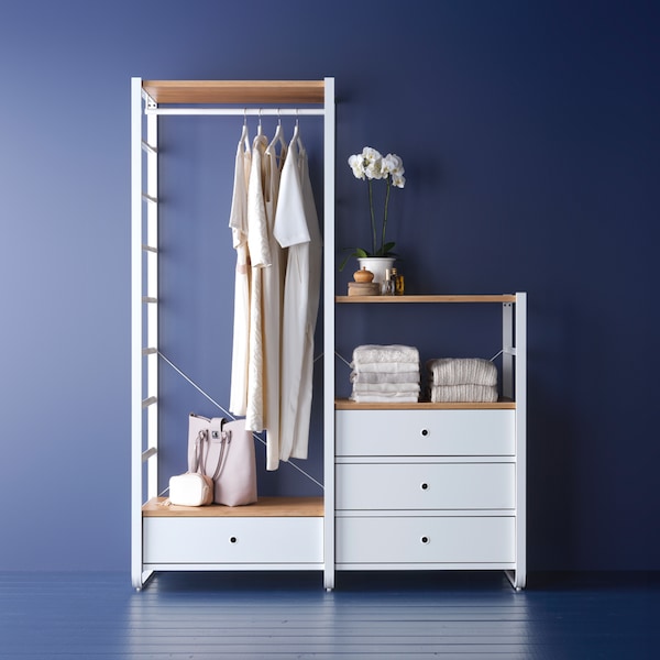 A white and bamboo ELVARLI storage combination against a dark blue wall with white dresses on hangers and tops neatly folded.