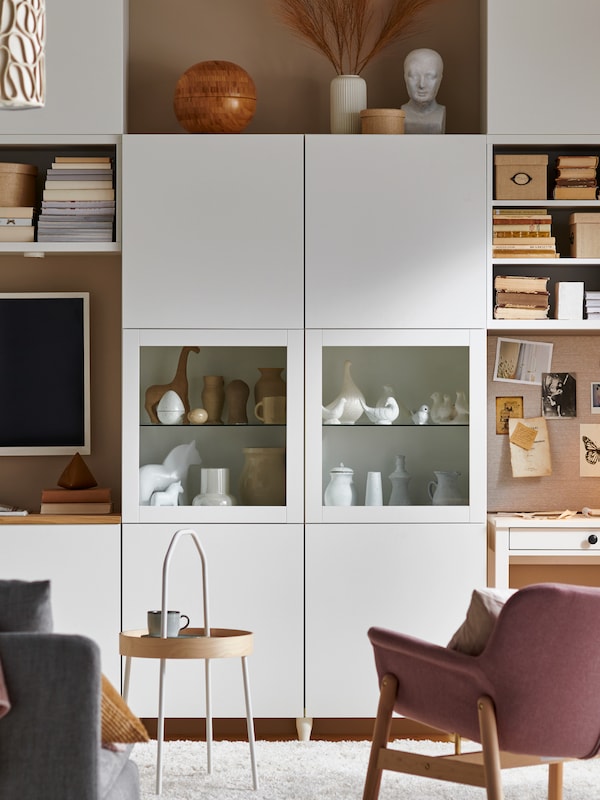 A white BESTÅ storage system showing decorative items, a TV on the wall, and a light brown-pink VEDBO armchair.