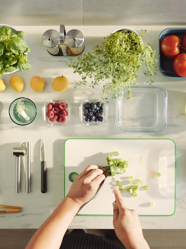 A white countertop with a person chopping green vegetables on it and fruit and veg in glass and china containers around them.