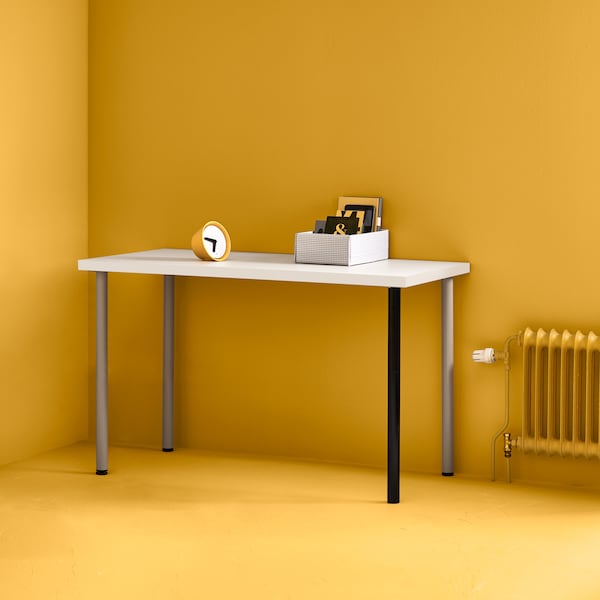 A white LINNMON table top with silver and black legs in the corner of a bright yellow room with a yellow radiator beside it.