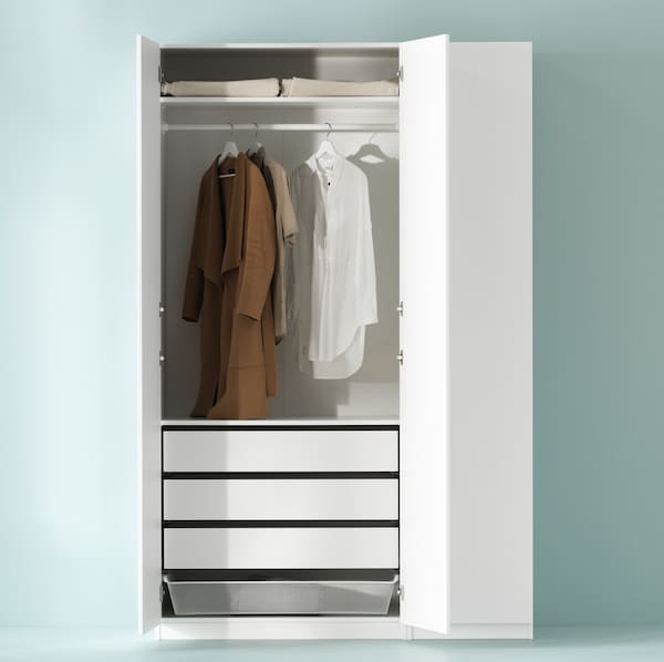 A white PAX wardrobe combination against a pale green wall with two doors open revealing three pieces of clothing on hangers.
