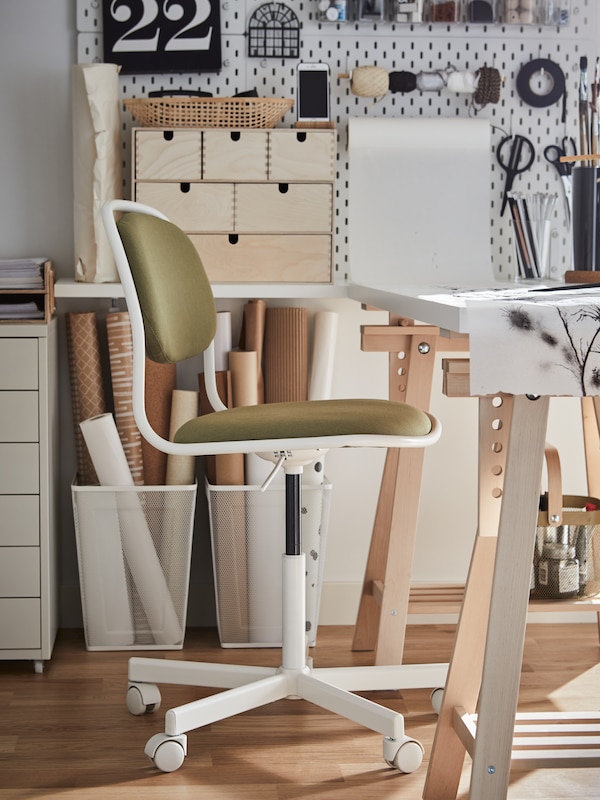 A white/yellow-green swivel chair, a white desk with wooden trestles, two white baskets with rolls of crafting paper.