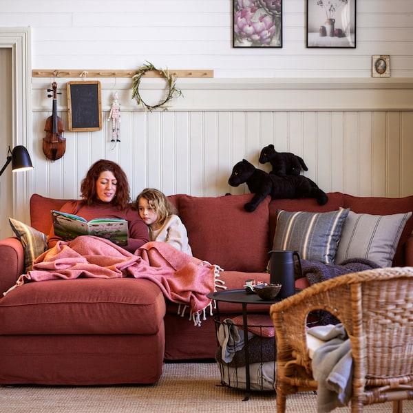 A woman and a girl are covered in a throw and read a book together on a light-red GRÖNLID three-seat sofa with chaise longue.